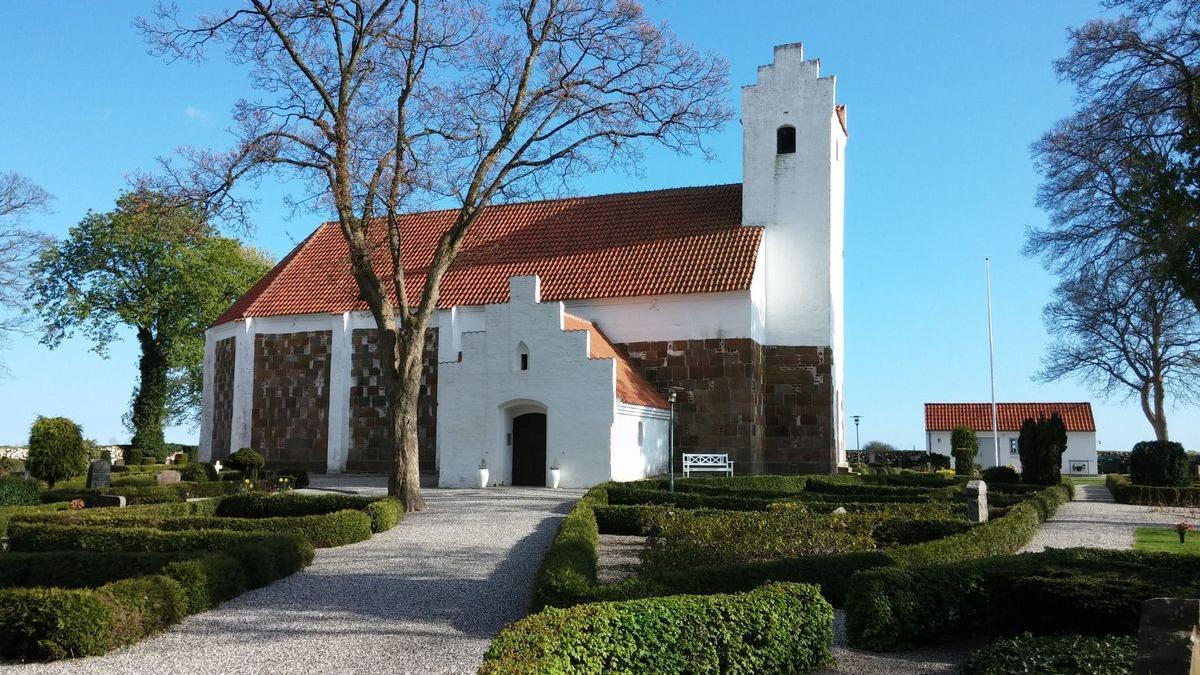 En hvid kirke med rødt tag og stenfacade omgivet af velplejede haver og træer under en blå himmel.