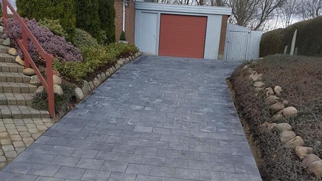 Nybelagt indkørsel med grå fliser og rød garageport, omgivet af buske og sten.