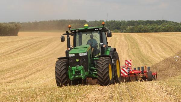 En grøn traktor pløjer en gylden mark under en skyet himmel.