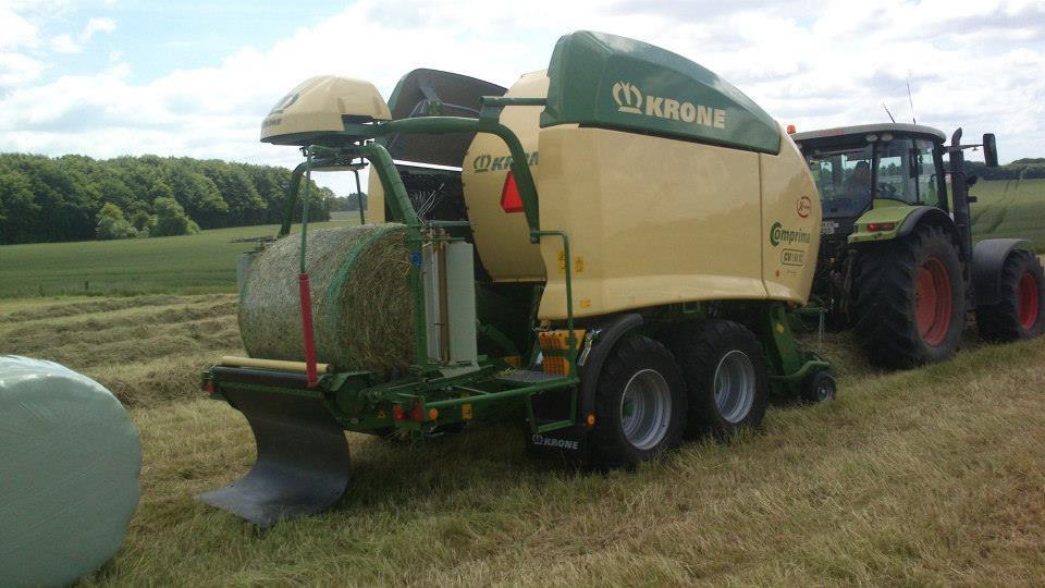 En stor høstmaskine fra Krogman, der pakker halmballer på en mark med en traktor i baggrunden.