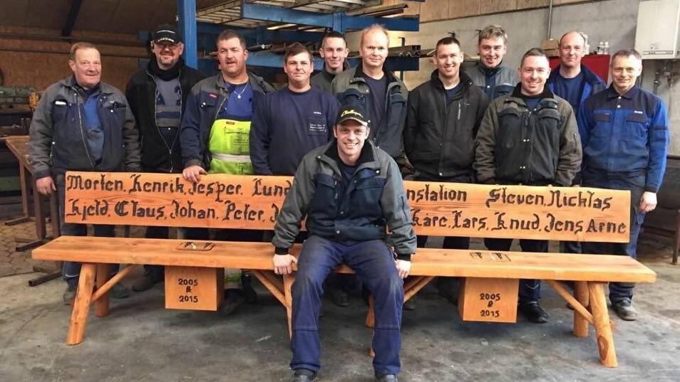 Gruppebillede af mænd, der står og sidder foran en træbænk med navne indgraveret.