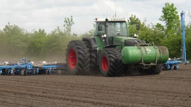 En stor traktor med grøn tank pløjer en mark under en skyet himmel.