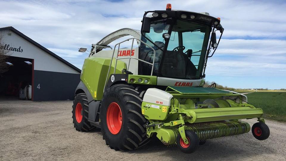 En Claas høstmaskine parkeret foran en lade med grønne marker i baggrunden.