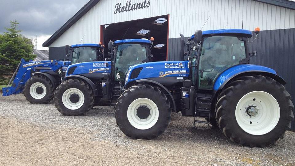 Tre blå traktorer parkeret foran en lade med skiltet "Hollands".