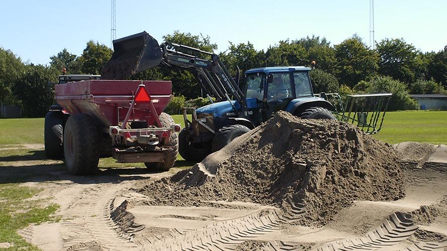 En traktor læsser sand fra en vogn på en græsplæne, omgivet af træer og en klar blå himmel.