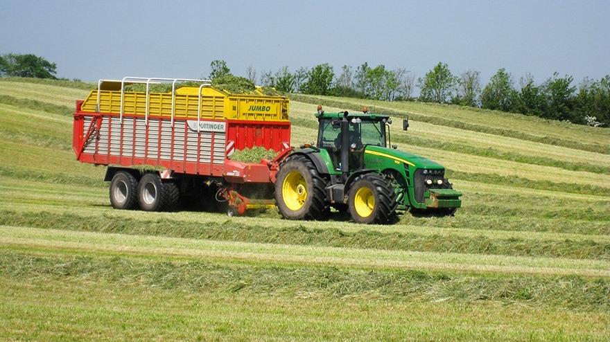 En grøn traktor trækker en stor vogn fyldt med hø på en mark under en blå himmel.