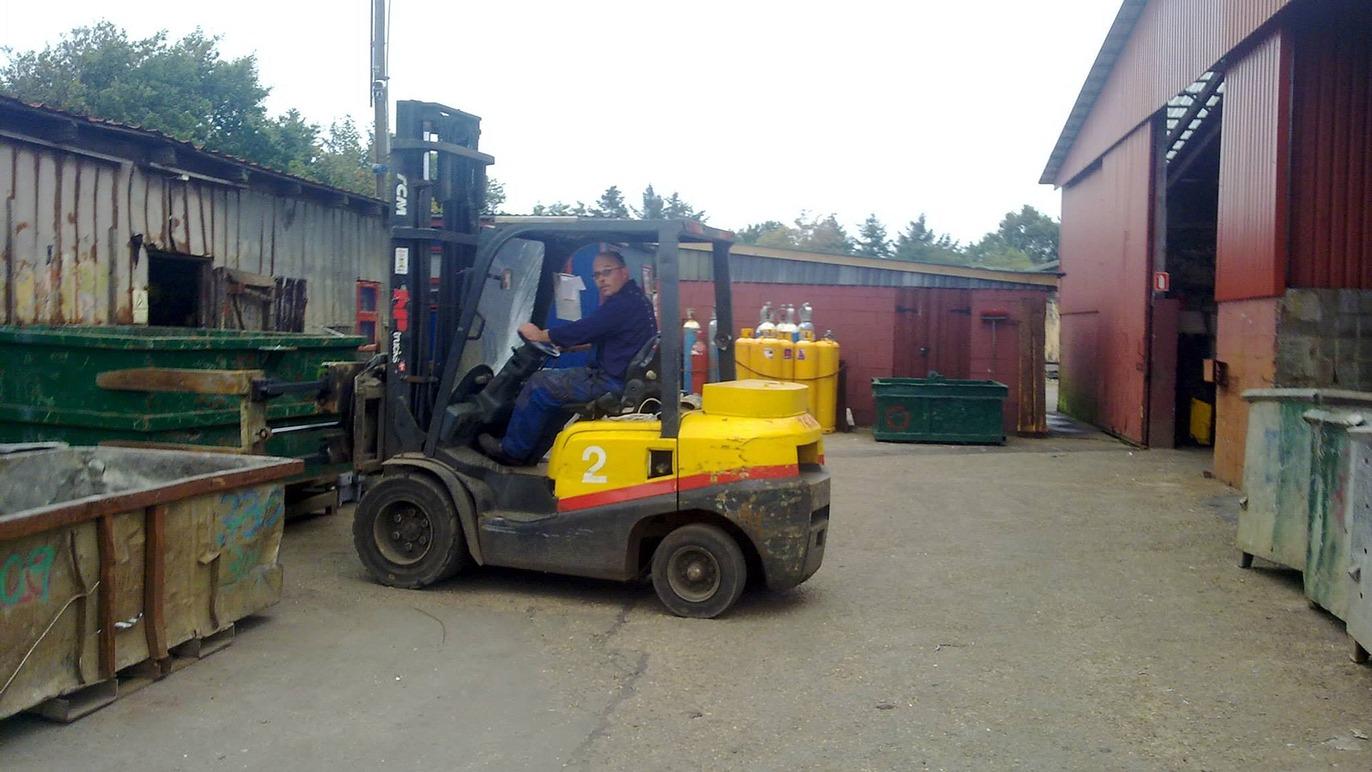 En mand kører en gaffeltruck med gule tanke i en gård med affaldscontainere og bygninger i baggrunden.