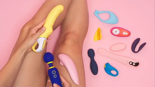 Hånd, der holder en gul vibrator, omgivet af forskellige sexlegetøj på en lyserød baggrund.