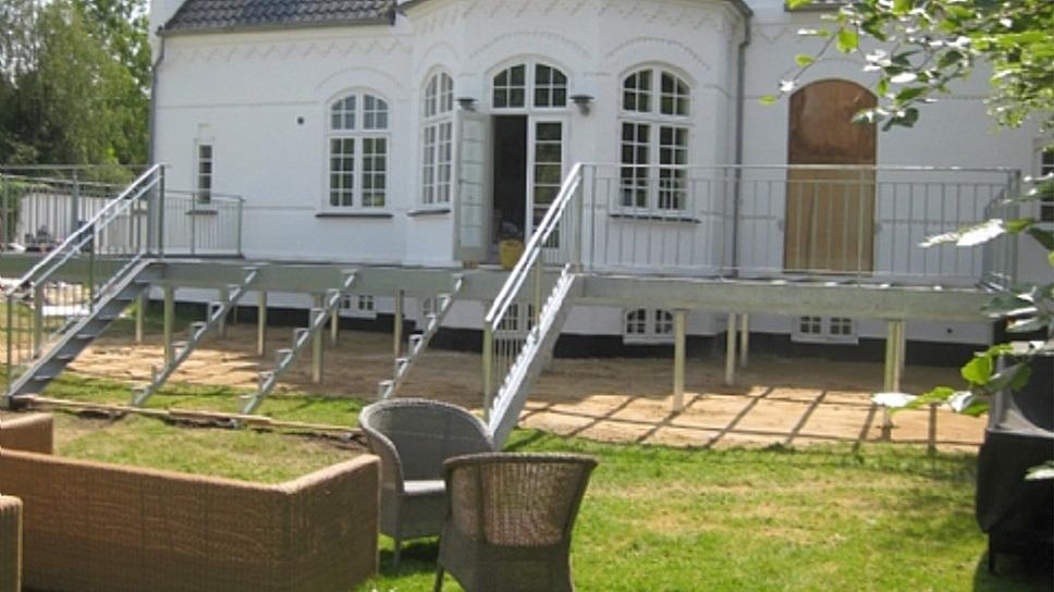 Hus med hvid facade, trapper og terrasse i haven, samt havemøbler i forgrunden.
