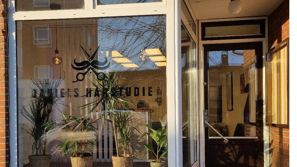 Frontal udsigt af en frisørsalon med planter og et skilt med teksten "Juliens Hårstudie" på vinduet.