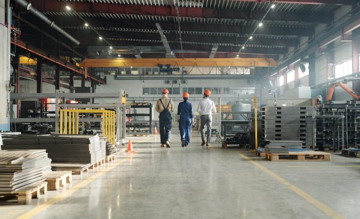 Tre arbejdere i sikkerhedshjelme går gennem en stor fabrikshal med metalplader og maskiner.