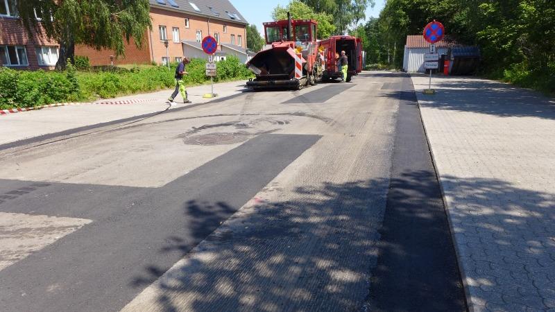 Vejarbejde med asfaltmaskine og arbejdere på en vej, omgivet af bygninger og trafikskilt.