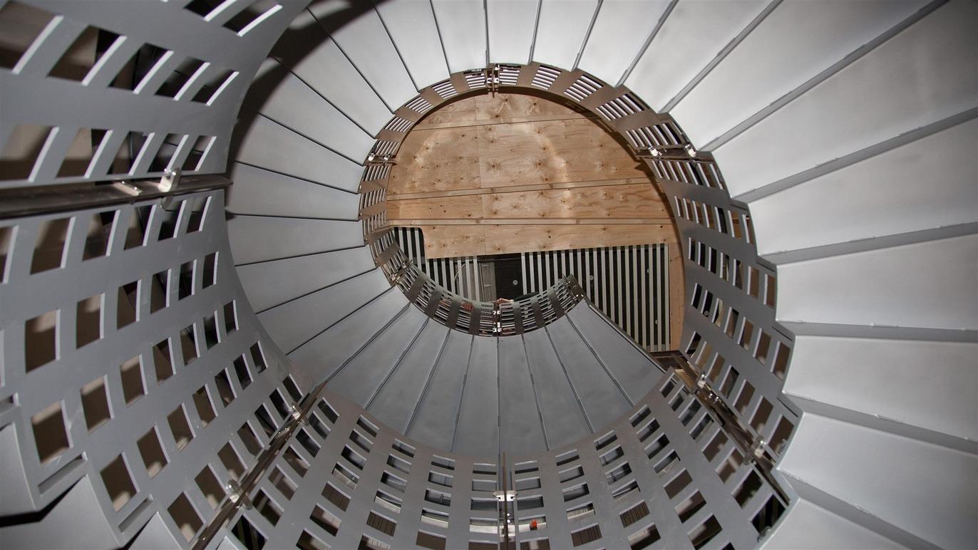 En spiraltrappe set oppefra med metalgelænder og træbund, der skaber en moderne og stilren atmosfære.