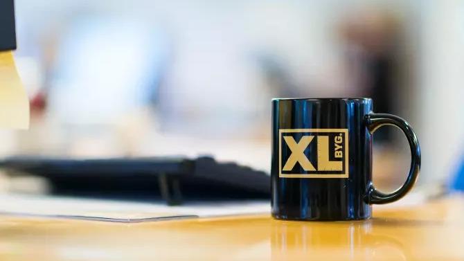 Sort krus med XL BYG-logo på et skrivebord ved siden af en computer og papirer.