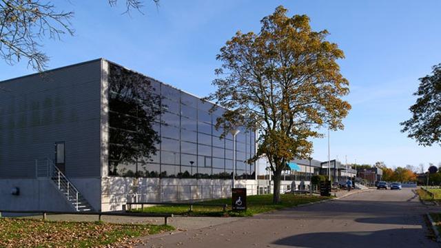 Moderne bygning med spejlende glasfacade og træer på en solrig dag. Vejen fører til parkeringspladsen.