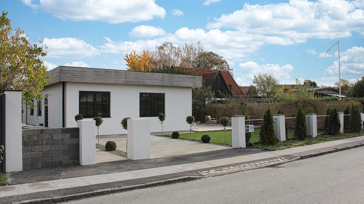Moderne hus med grå tag, hvid facade og velplejet have, beliggende ved en stille vej.