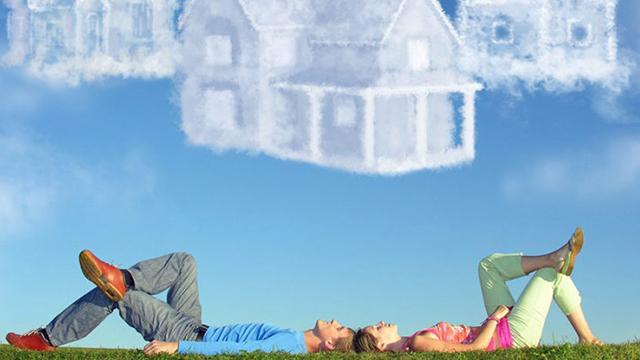 To personer ligger på græsset og kigger op på skyer, der ligner huse i en blå himmel.