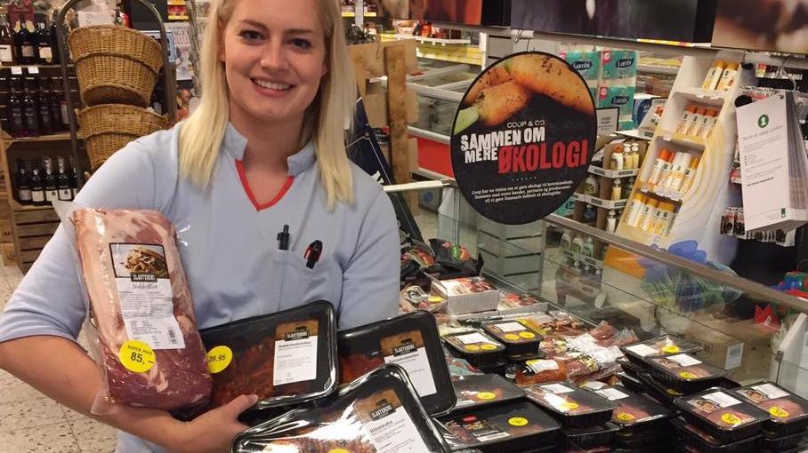En smilende kvinde holder kødprodukter i en butik med økologiske varer i baggrunden.