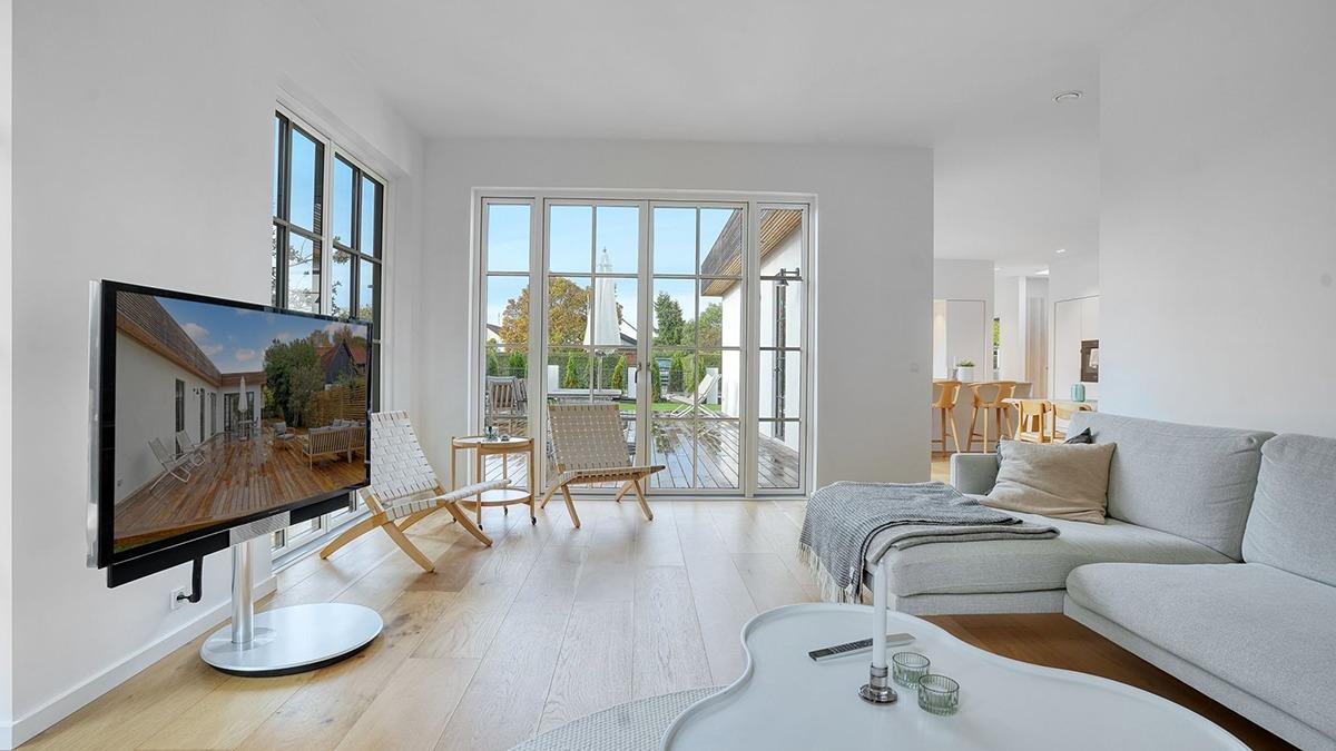 Moderne stue med grå sofa, fladskærms-tv og store vinduer, der giver udsigt til en terrasse.