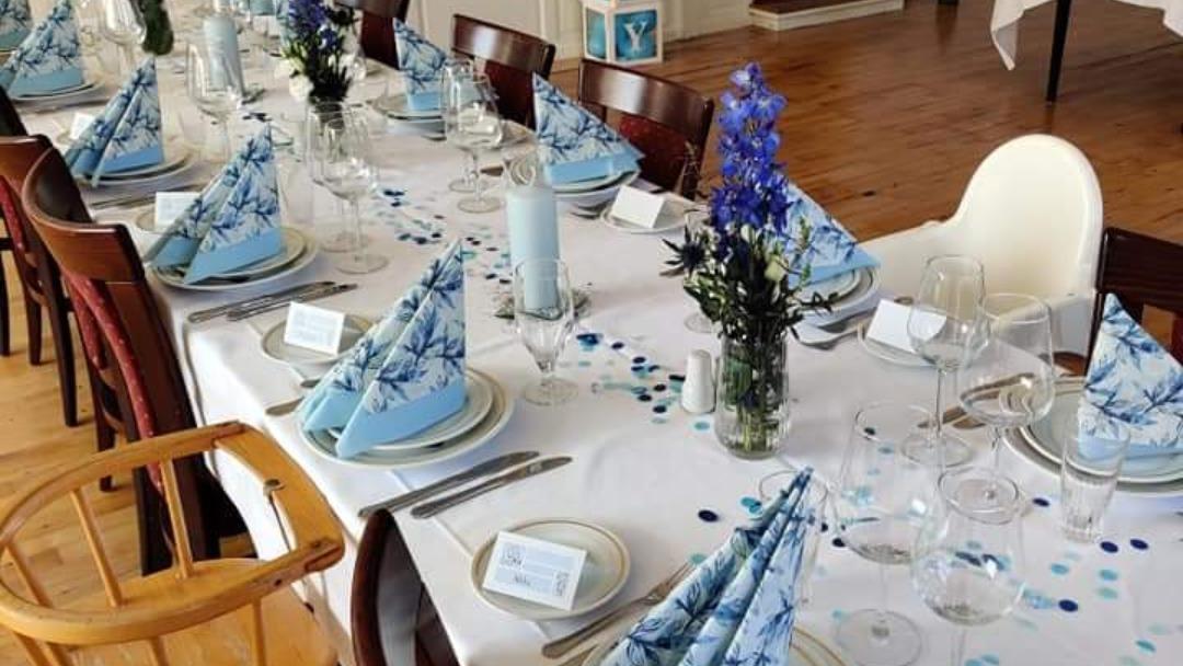 Elegant dækket bord med blå servietter, blomster og glas til festlig middag.