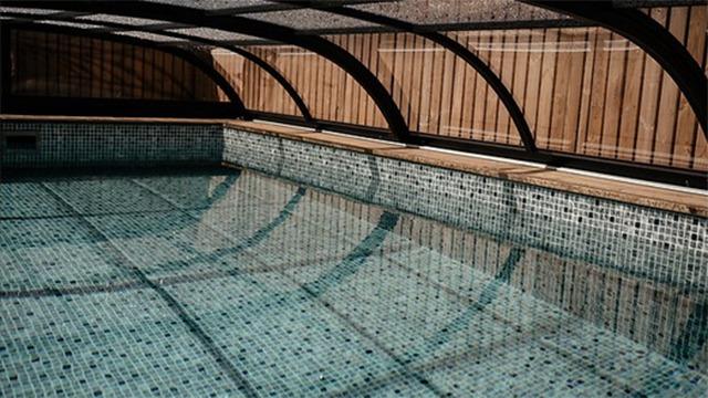Indendørs swimmingpool med mosaikfliser og et buet glasloft, der reflekterer vandets overflade.