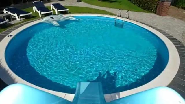 Blå swimmingpool med rutsjebane og liggestole i en have. Solskin og grøn græsplæne i baggrunden.