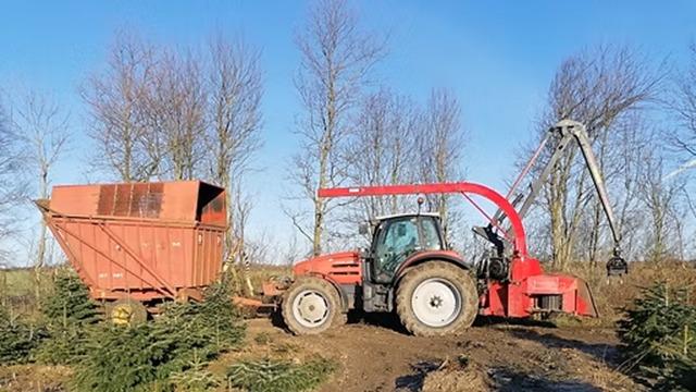 Rød traktor med en stor maskine, der arbejder i en skov med træer og en vogn bagpå.