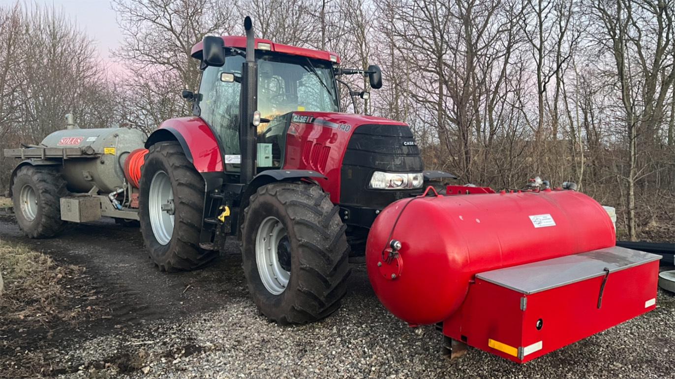 Rød traktor med gyllevogn på en grusvej omgivet af træer i skumringen.