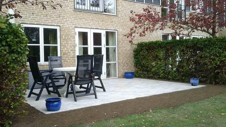 En hyggelig terrasse med et bord og stole, omgivet af grønne buske og blomsterkrukker.