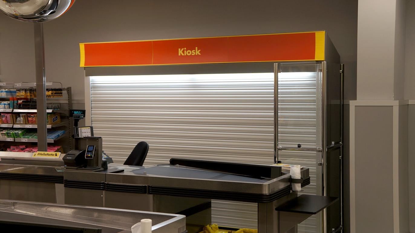 Kiosk med lukket rulleport og orange skilt i en butik, med en kasseapparat og stole synlige.