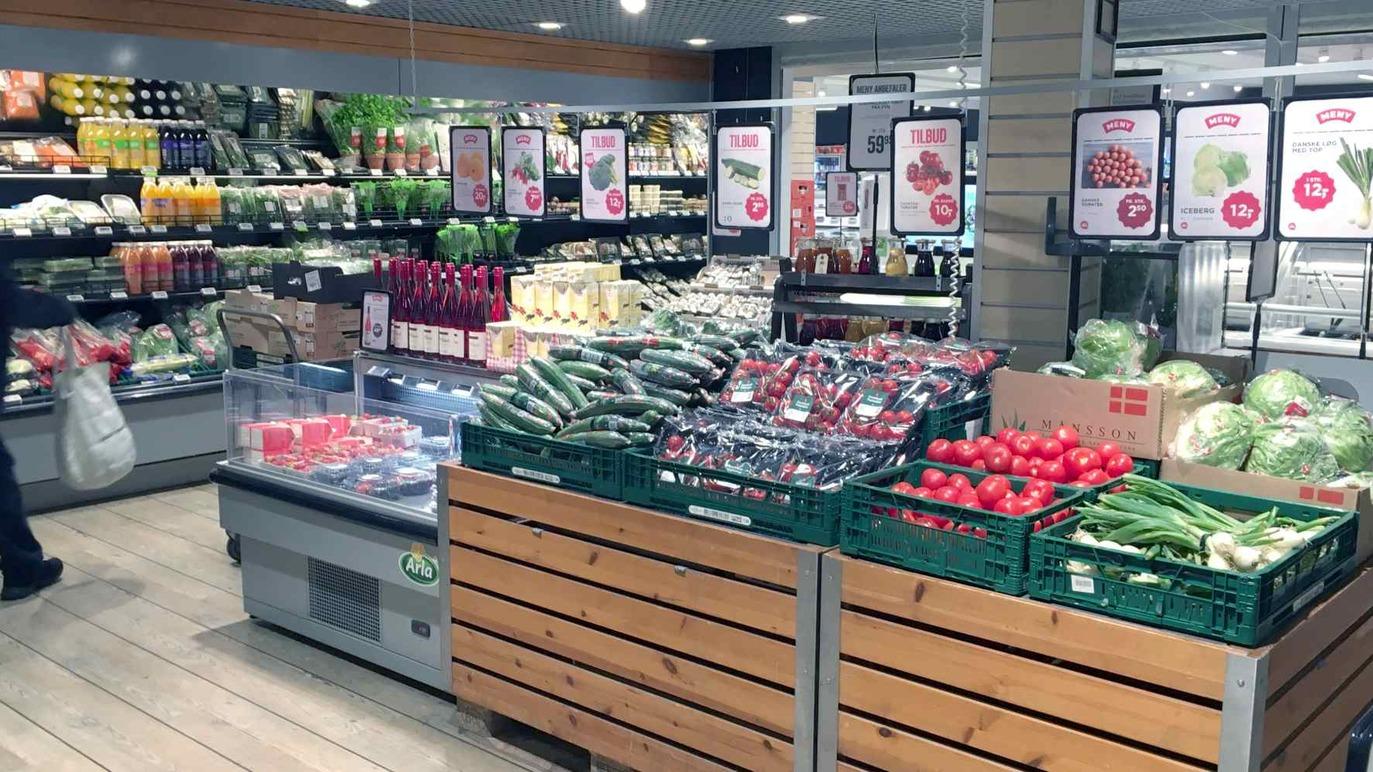 Frisk frugt og grøntsager præsenteret i en supermarked med køleskabe og skilte.