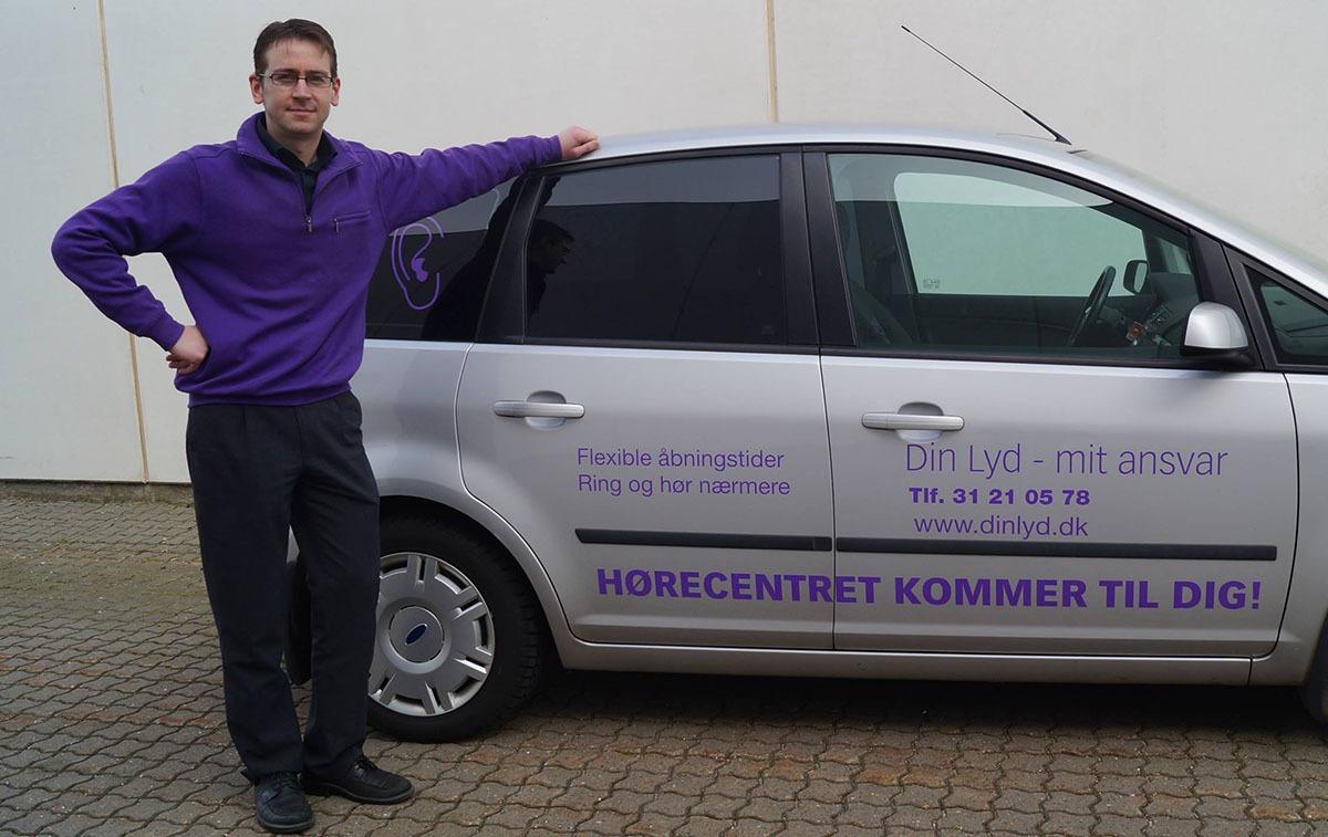 En mand står ved siden af en sølvfarvet bil med teksten "Horecentret kommer til dig!" på siden.