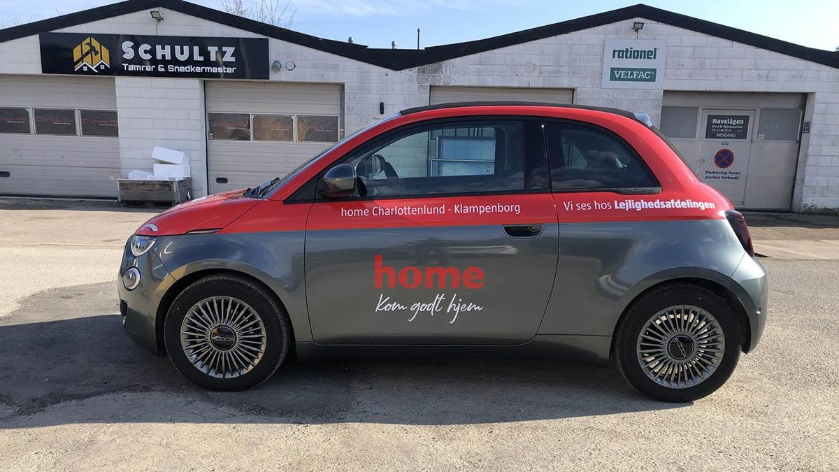 En grå bil med rød tag og reklame for "home" parkeret foran en bygning med garager.