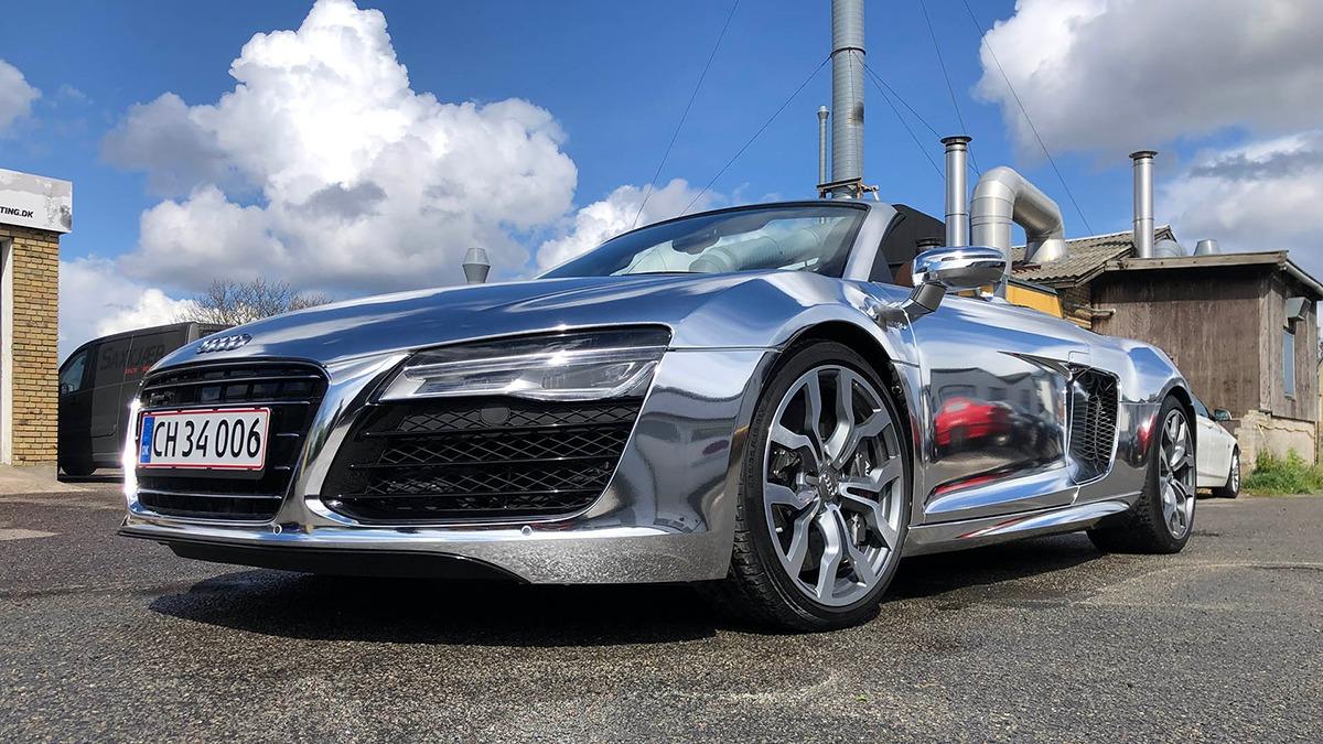 En skinnende sølvfarvet Audi R8 sportsvogn parkeret på en vej med skyer i baggrunden.