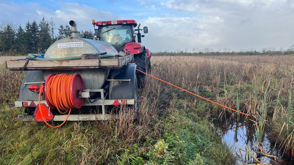 Traktor med slange, der arbejder i en mark med højt græs og vandpytter under en skyet himmel.