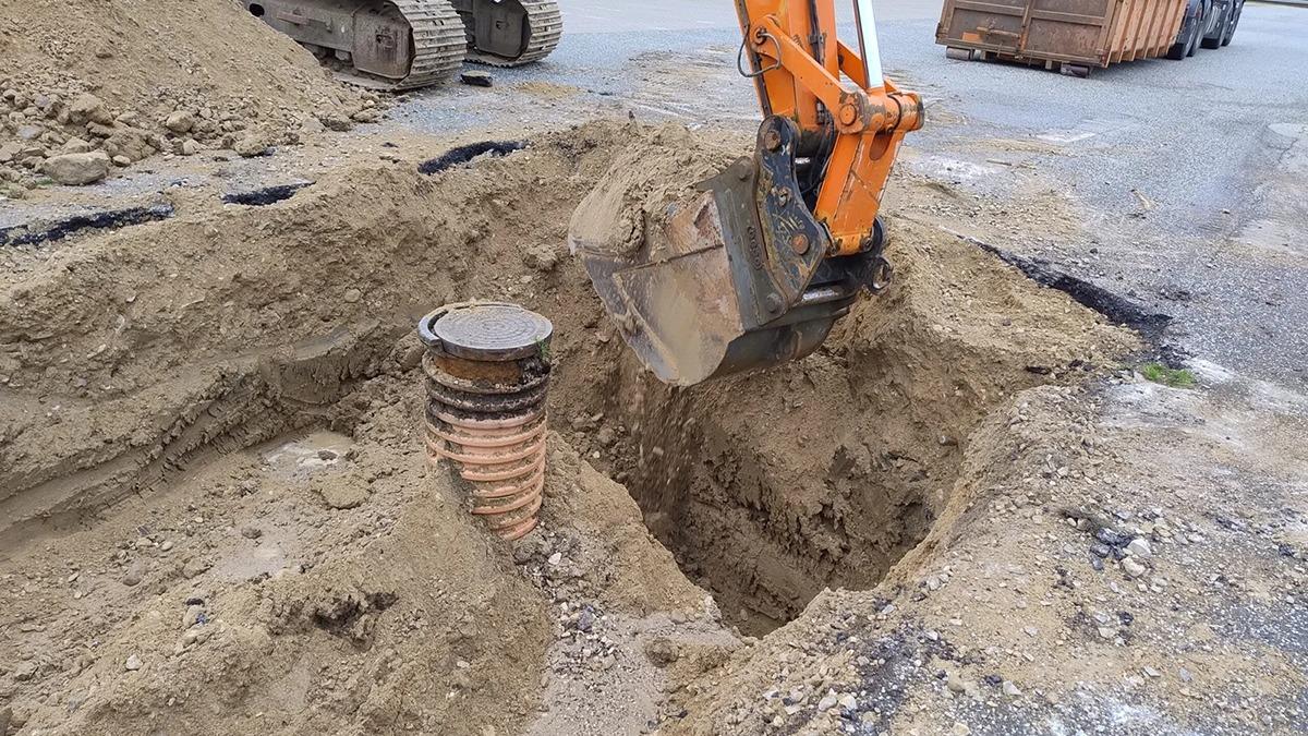 Gravemaskine arbejder i en stor grav, hvor en brønd er synlig i sandet.