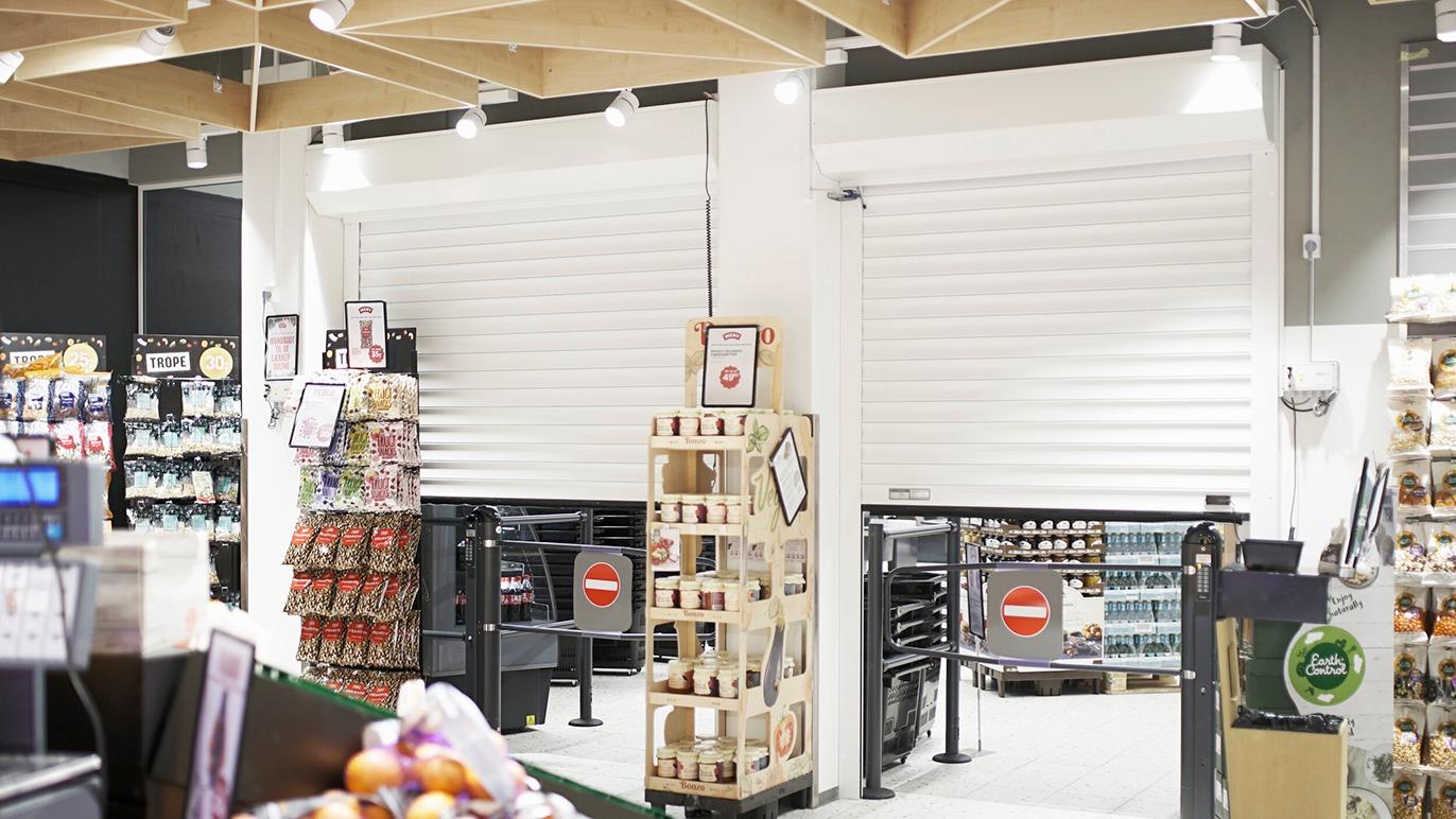 Indgang til en butik med rulleporte og hylder fyldt med varer i baggrunden.