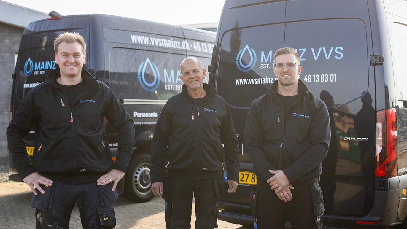Tre VVS-teknikere står foran deres varevogne med logoet fra Manz VVS, iført sorte jakker og arbejdstøj.