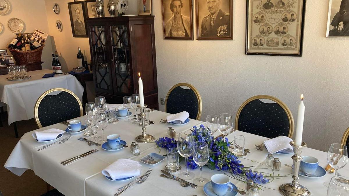Elegant dækket bord med blå tallerkener, glas og lysestager i en hyggelig spisestue med vintage billeder på væggene.