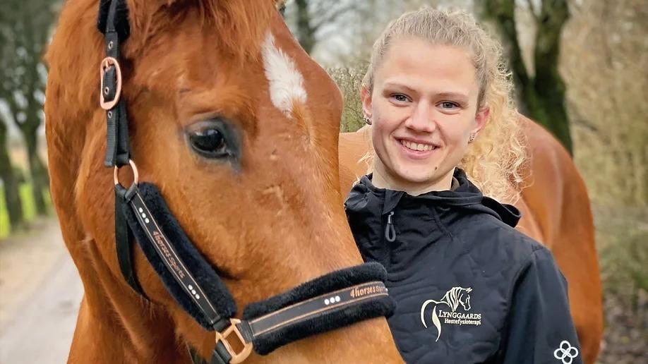 En ung kvinde smiler ved siden af en brun hest med en hvid stjerne i panden, begge står i naturen.