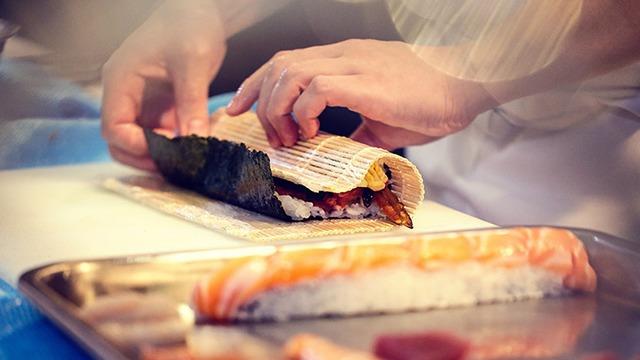 Hænder der ruller sushi med tang og fyld på en skæreplade i et køkken.