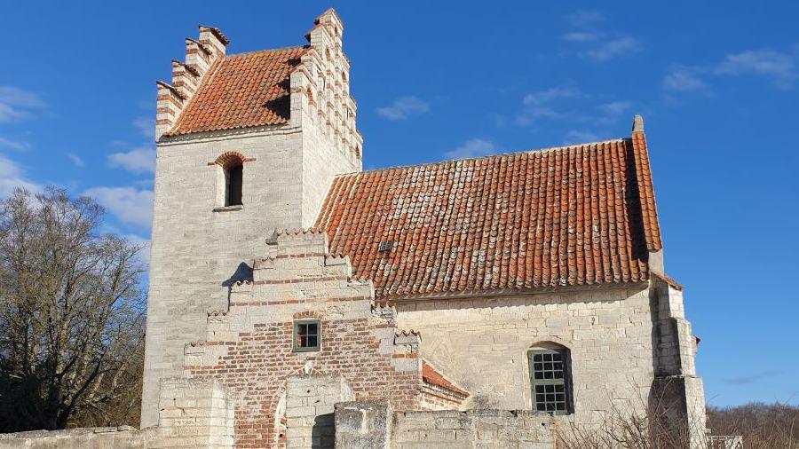 En gammel kirke med rød teglsten og en karakteristisk tårn, omgivet af blå himmel og træer.