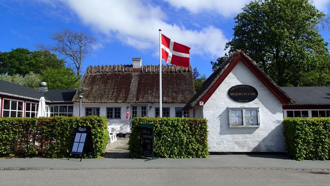 En hyggelig bygning med stråtag og dansk flag, omgivet af grønne buske og en klar blå himmel.