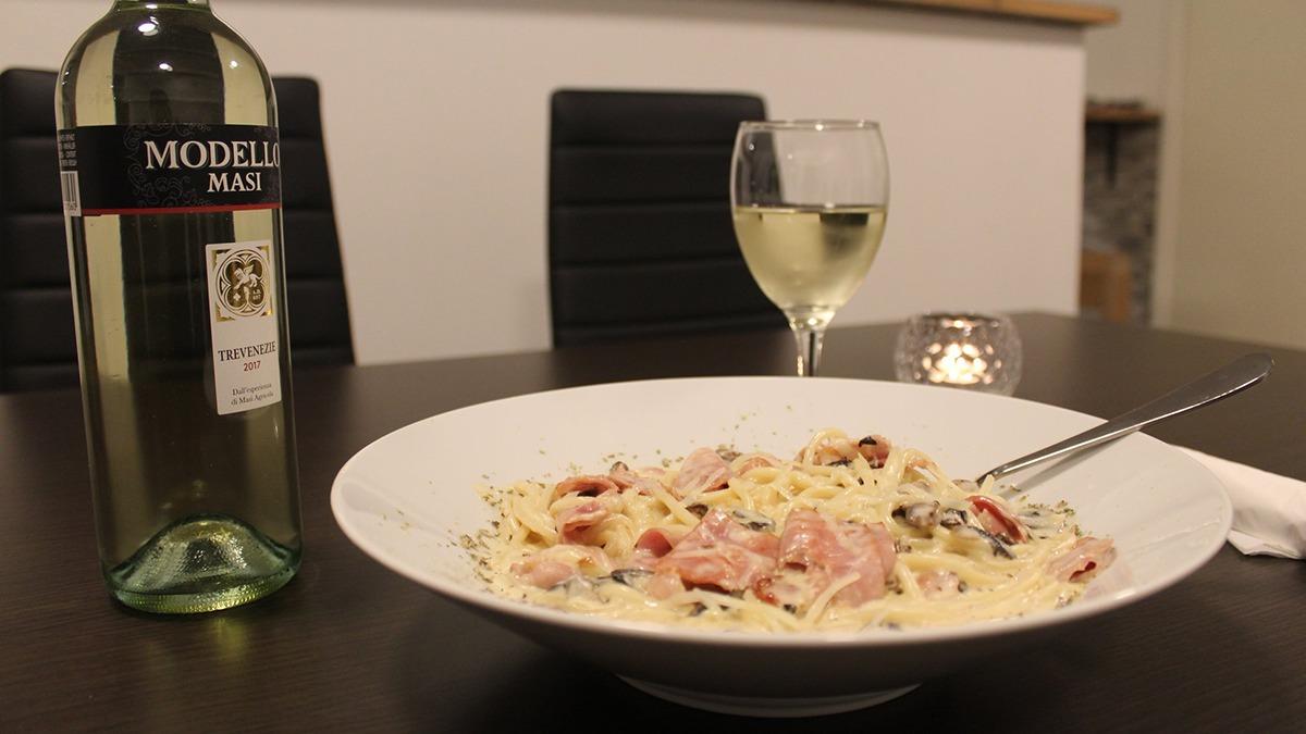 En skål med pasta i flødesauce og skinke, en flaske hvidvin og et glas vin på et bord.