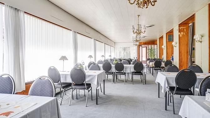 En lys restaurant med borde dækket med hvide duge og stole, store vinduer og elegant belysning.