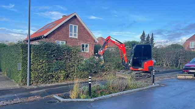 Gravemaskine arbejder på at fjerne buske foran et rødt hus på en vej med biler og blå himmel.