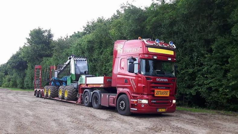En rød Scania lastbil med en grøn maskine og en trailer parkeret på en grusvej omgivet af træer.