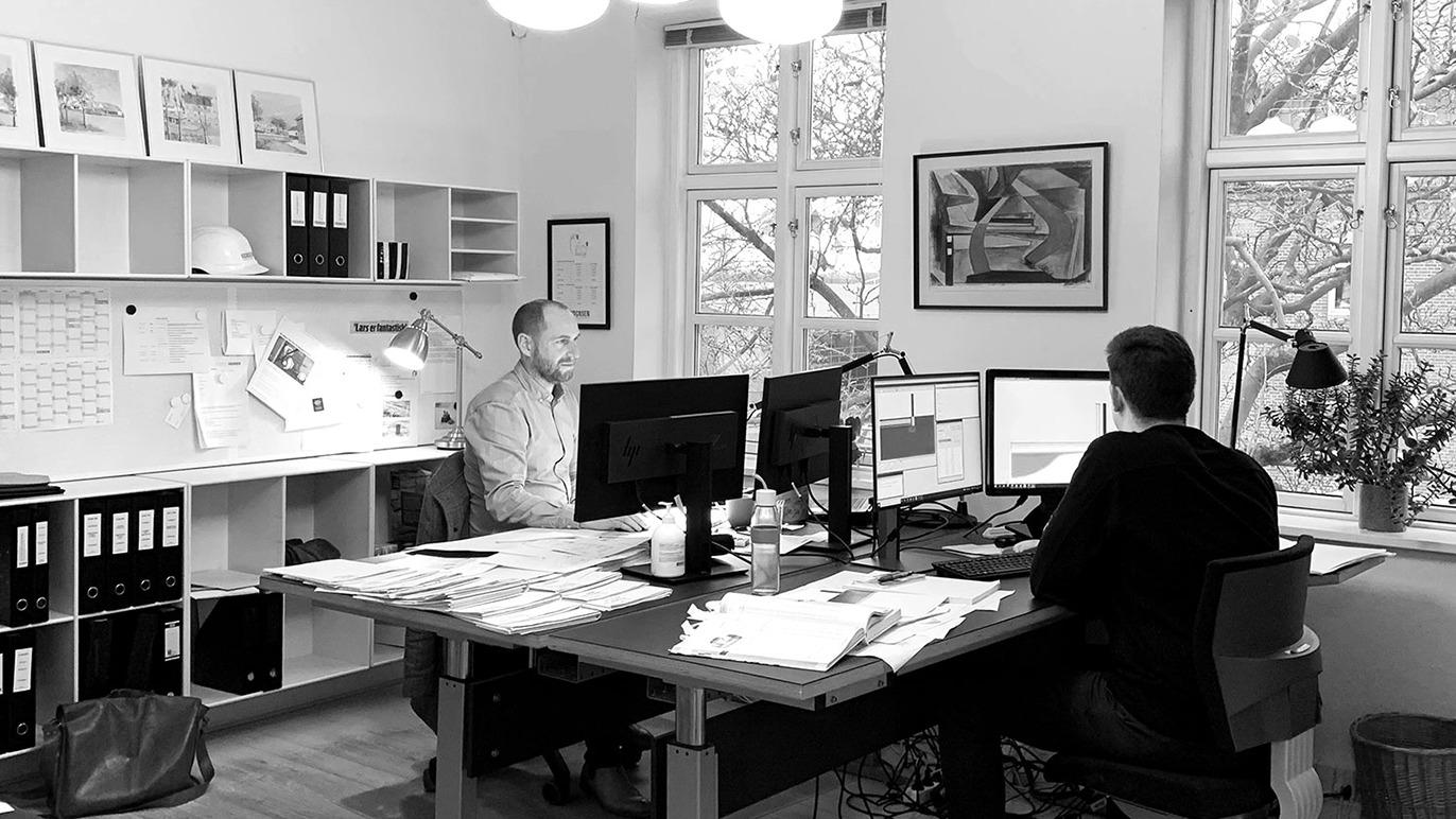 To personer arbejder ved skriveborde i et lyst kontor med store vinduer og planter.