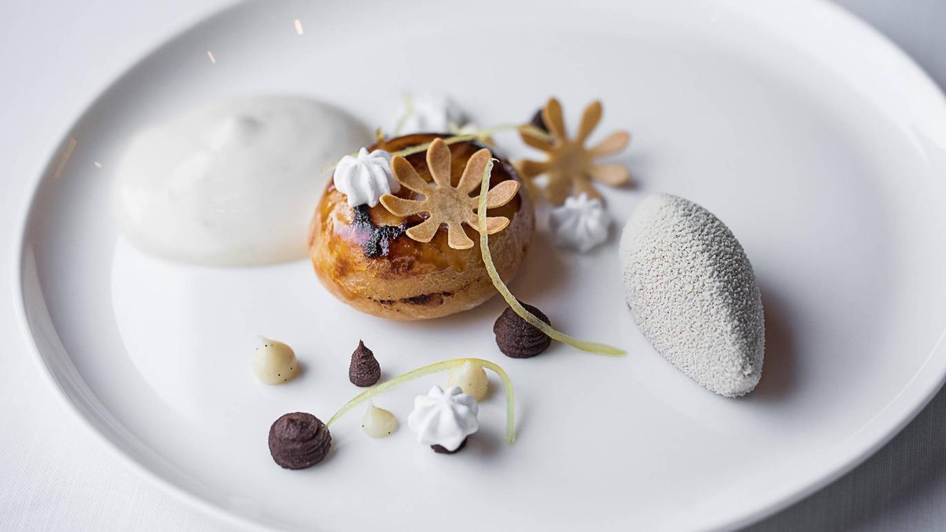 En elegant dessert anrettet på en hvid tallerken med blomster, skum og chokolade.