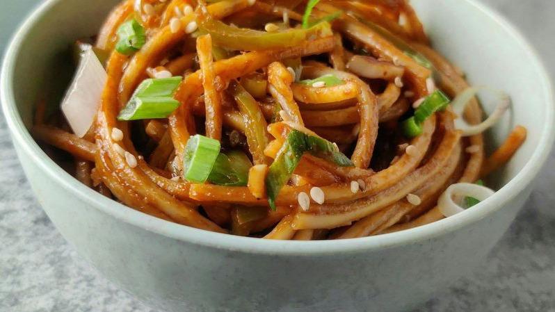 En skål med nudler, grøntsager og sesamfrø, serveret med forårsløg og en lækker sauce.